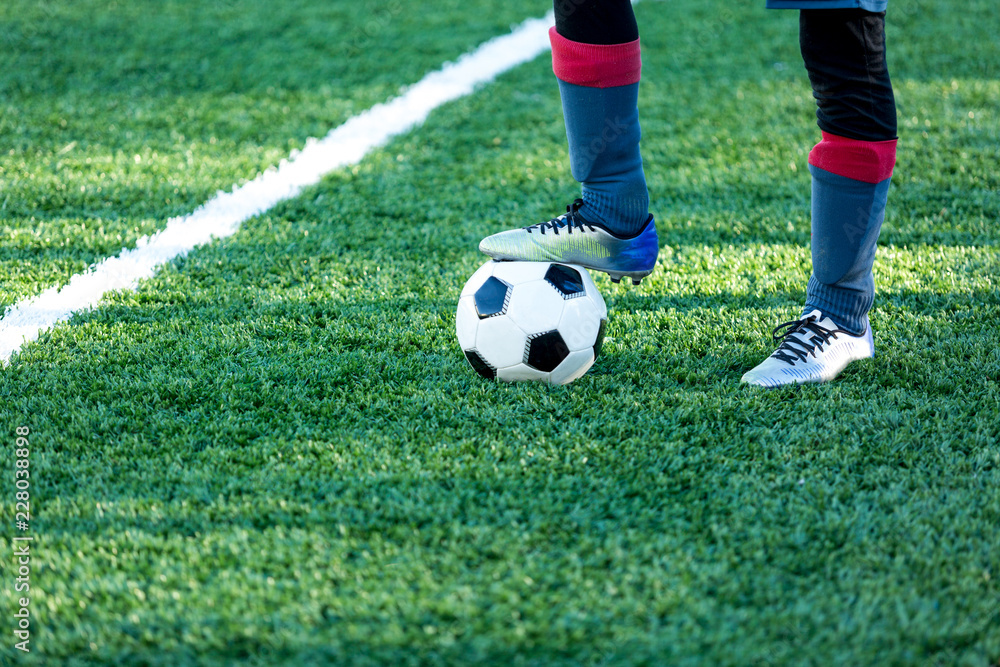 Obraz premium classical black and white football ball on the green grass of the field and leg in sports shoes on it. Soccer game, training, hobby concept. with copy space 