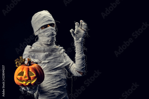 Obraz na plátně Studio shot portrait  of young man in costume  dressed as a halloween  cosplay of scary mummy pose like a hold jack'o pumpkin clamber acting on isolated black background