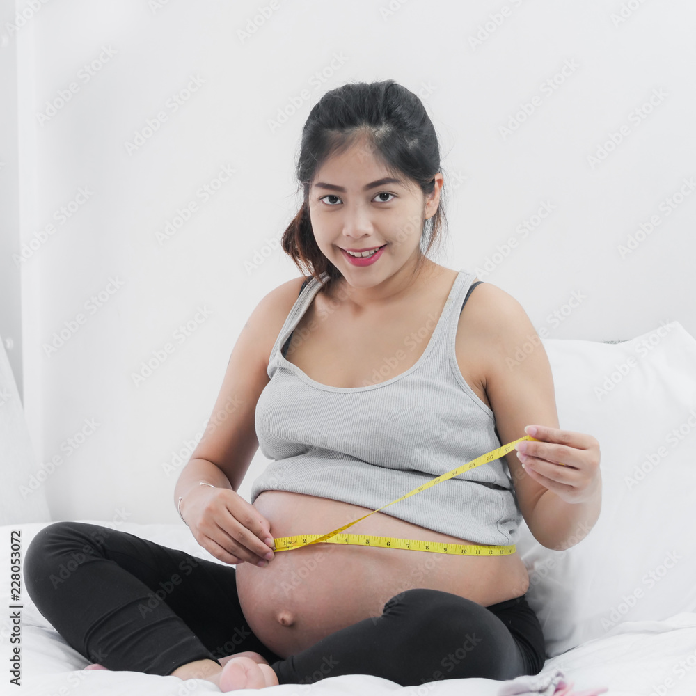 Happy Asian pregnant woman using measuring tape with her belly, lifestyle concept.
