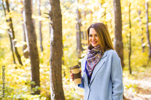 Autumn, nature, people concept - young brunette woman in a blue coat standing in the park with a cup of coffee