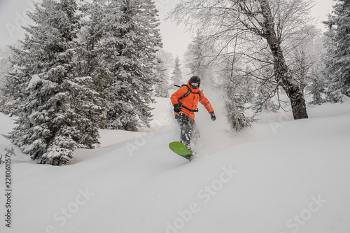 Man riding down the hill on the green snowboard between trees in the mountain resort