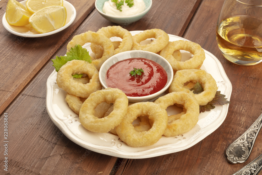 A closeup photo of squid rings with a tomato sauce and Mayonnaise, lemon wedges, and white wine, on a dark rustic wooden background
