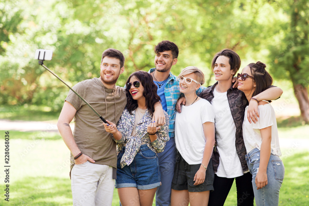 friendship, leisure and technology concept - group of happy smiling friends taking picture by selfie stick at summer park
