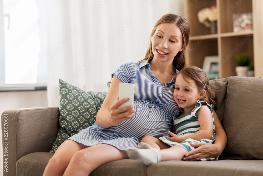 pregnancy, people and family concept - happy pregnant mother and little daughter with smartphone at home