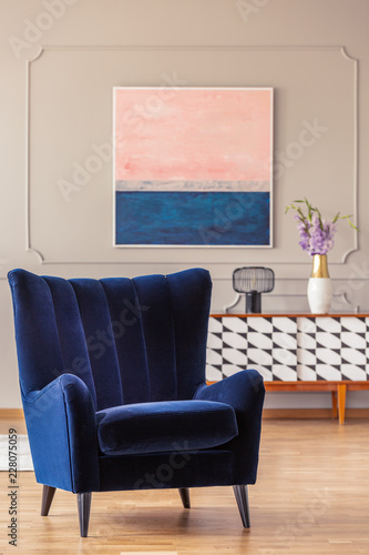 Retro, navy blue armchair in an elegant living room interior with an abstract painting on a wall with molding