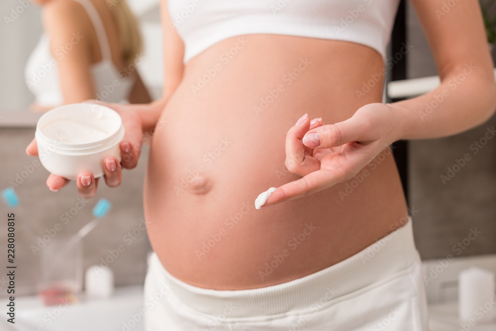 cropped shot of pregnant woman with cream near belly to avoid stretch marks in bathroom