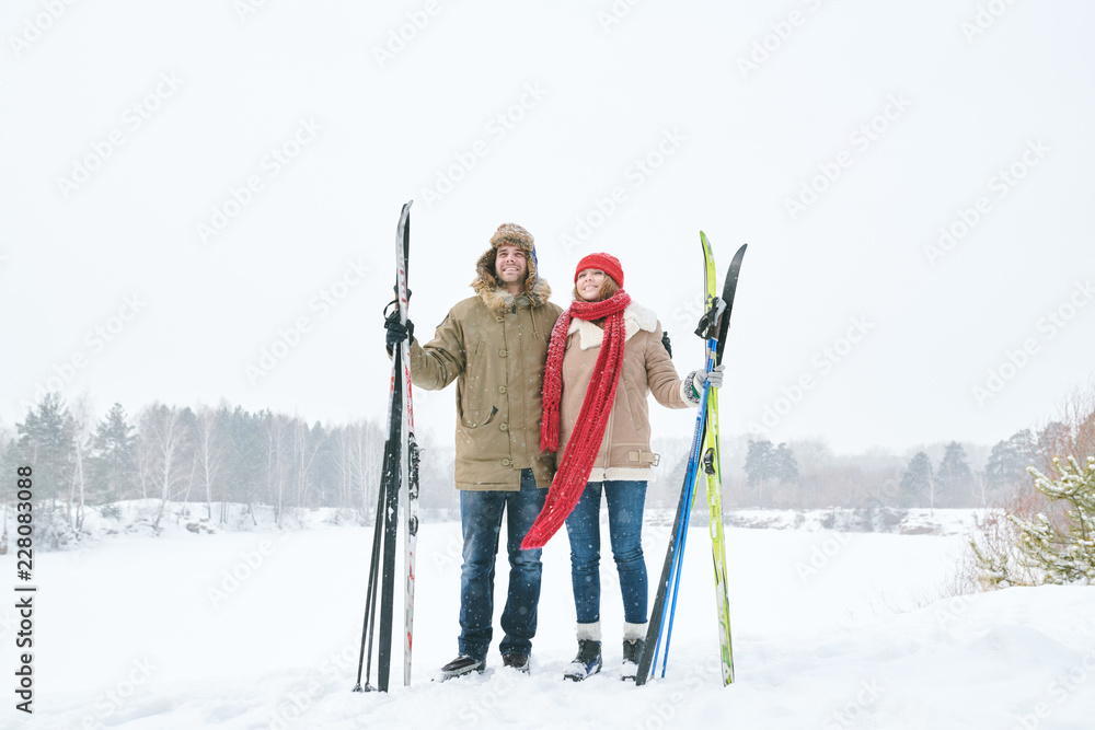 Full length portrait of loving young couple embracing and smiling happily while taking break during skiing trip and posing against beautiful winter landscape, copy space