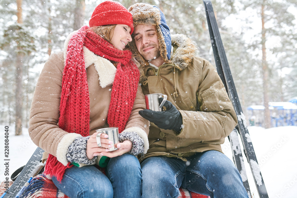 Obraz premium Portrait of loving young couple cuddling tenderly and enjoying hot cocoa during date in beautiful winter forest, copy space