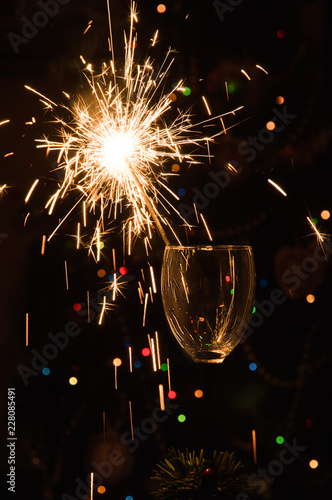 Macro, Close up. Вurning sparkler stands in a glass. Dark background with defocused multi-colored lights of garland.