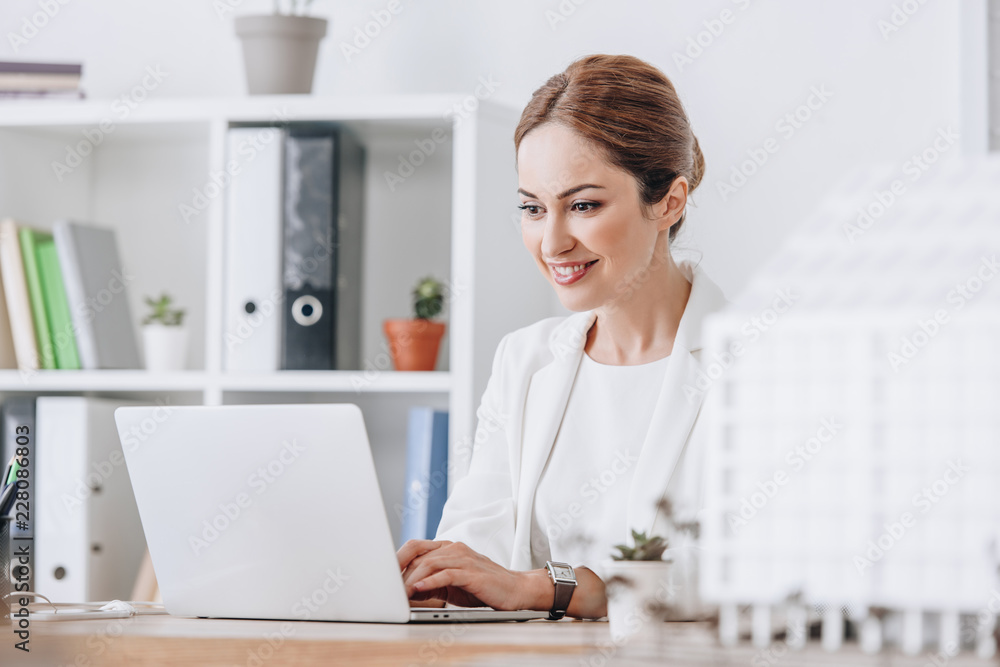 smiling female architect working with laptop at workplace with house model, selective focus smiling female architect working with laptop at workplace with house model, selective focus