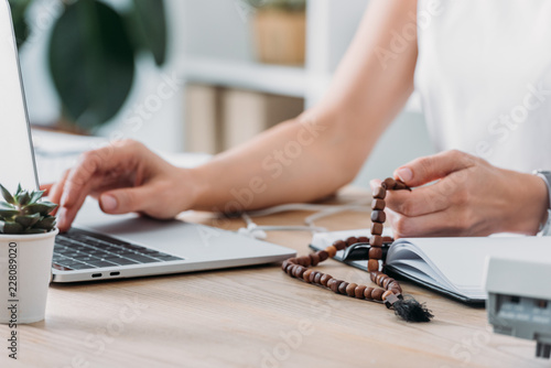 Photography cropped view of businesswoman using laptop and holding rosary beads at workplace