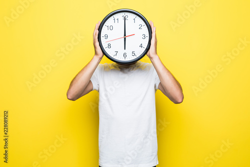 Young man holding big clock covering his face on yellow background