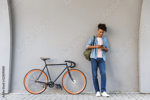 Happy young african guy walking outdoors with bicycle chatting by mobile phone.