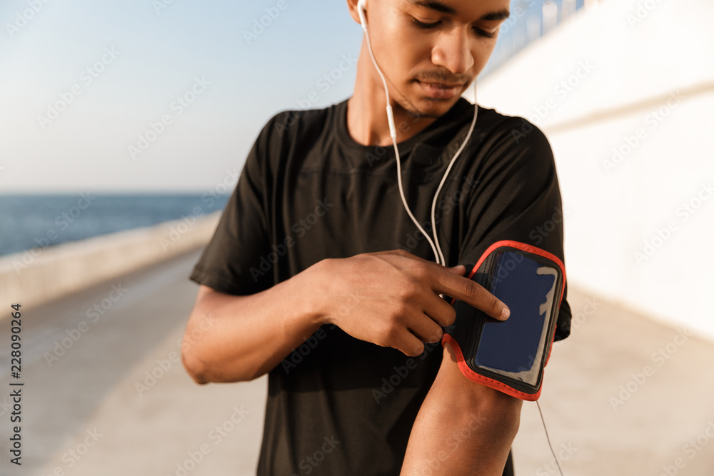 Fototapeta premium Guy sportsman outdoors on the beach listening music with earphones using phone.