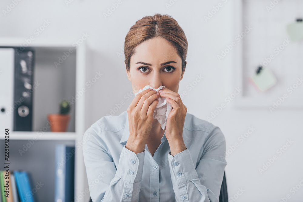 sick businesswoman with paper napkin having runny nose and looking at camera at office sick businesswoman with paper napkin having runny nose and looking at camera at office