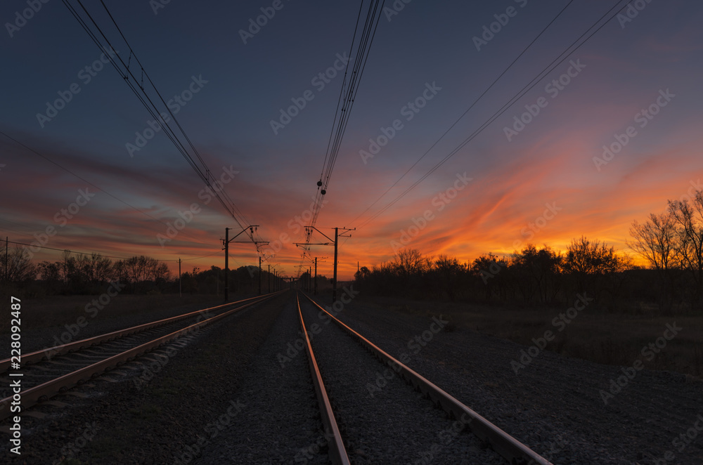 Sunset light reflects on the rails that running away to the horizon. Dark railway infrastructure at sunset. View of railroad going straight away to sun. The colorful sky with clouds at dramatic sundow