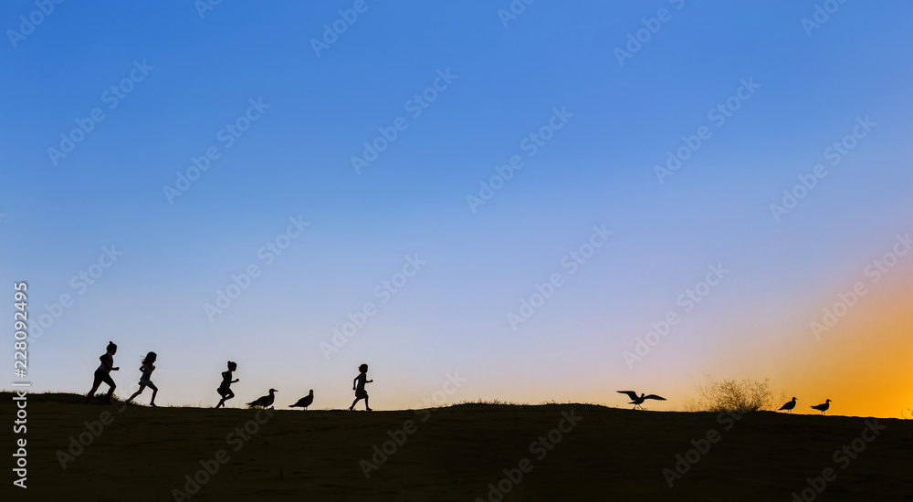 Silhouettes of four running kids and seagulls on the horizon on sunset. Sky is blue, sun sets in the right corner. Lots of empty space, copy space, for design, meme, background, wallpaper, poster, ad.