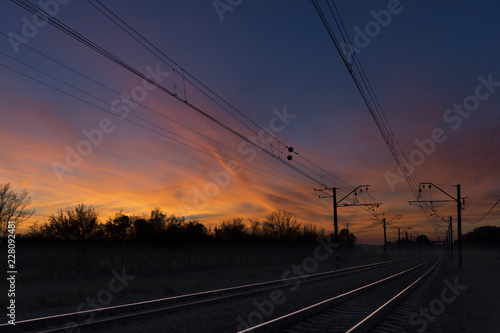 Dark silhouettes railway infrastructure against background of colorful dramatic sunset. View of railroad going straight away to sun and beyond the horizon. Transportation and travel concept.