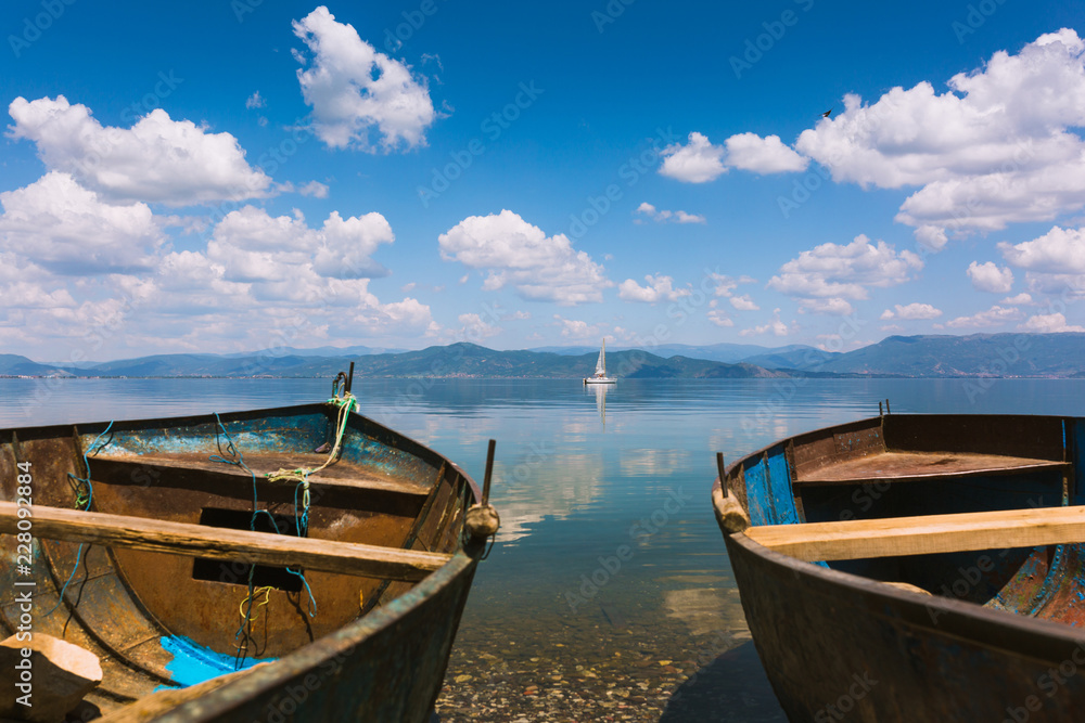 Two old fishing boats and a sailing ship in the distance. Blue lake with stones near the shore and clouds reflecting in the water. Mountains in the distance, also reflecting in the water. Sunny day.