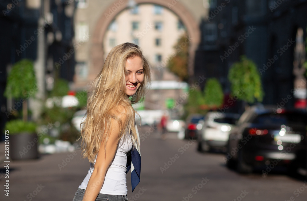Smiling blonde woman with long hair wearing shawl and white shirt  posing with sun light at the street
