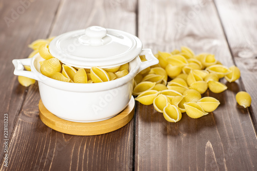 Lot of whole light raw yellow pasta conchiglie variety in a ceramic stewpan on brown wood