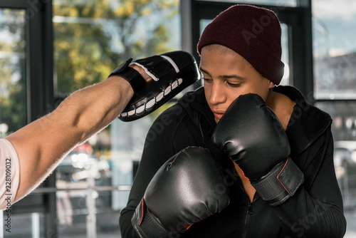 selective focus of african american boxer in boxing gloves practicing with trainer at gym