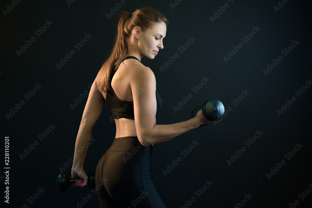 Obraz premium beautiful young girl training with dumbbell shaking her biceps on a black background