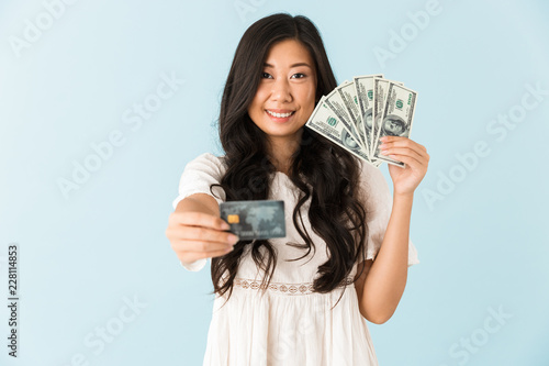 Happy asian beautiful woman isolated over blue background holding credit card and money.