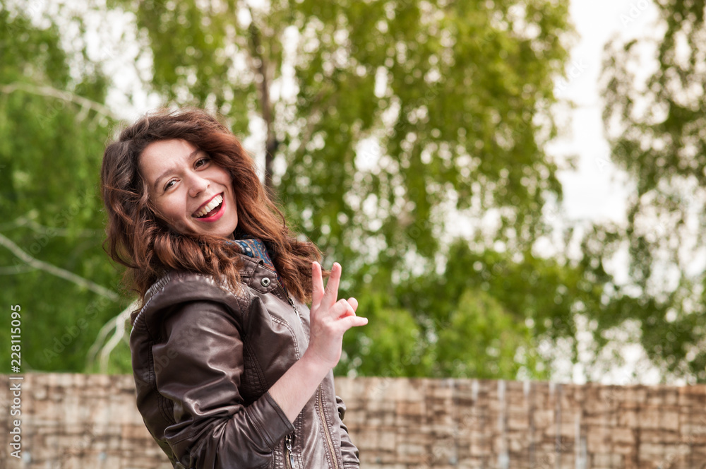 Cheerful with a broad smile a girl showing a peace sign in a leather jacket against a background of green foliage Cheerful with a broad smile a girl showing a peace sign in a leather jacket against a background of green foliage
