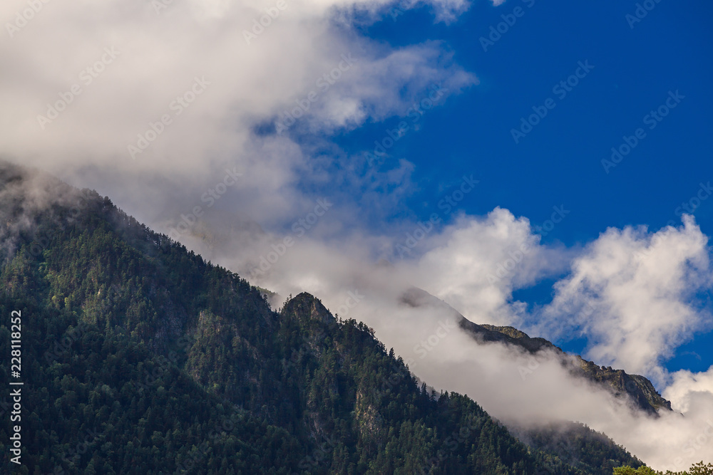 Clouds over the tops of the rocky mountains overgrown with trees. Photographed in the Caucasus, Russia.