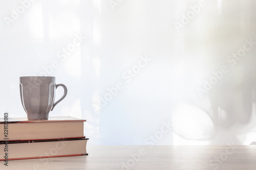 Photo of a white desk with thick books and a gray cup of hot tea on a background of sun-lit curtains. Copy space