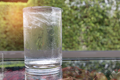 Cool drinking water in glass with ice inside on glass table with dry flower below, as natural in the morning with sunrise background. Drink concept.