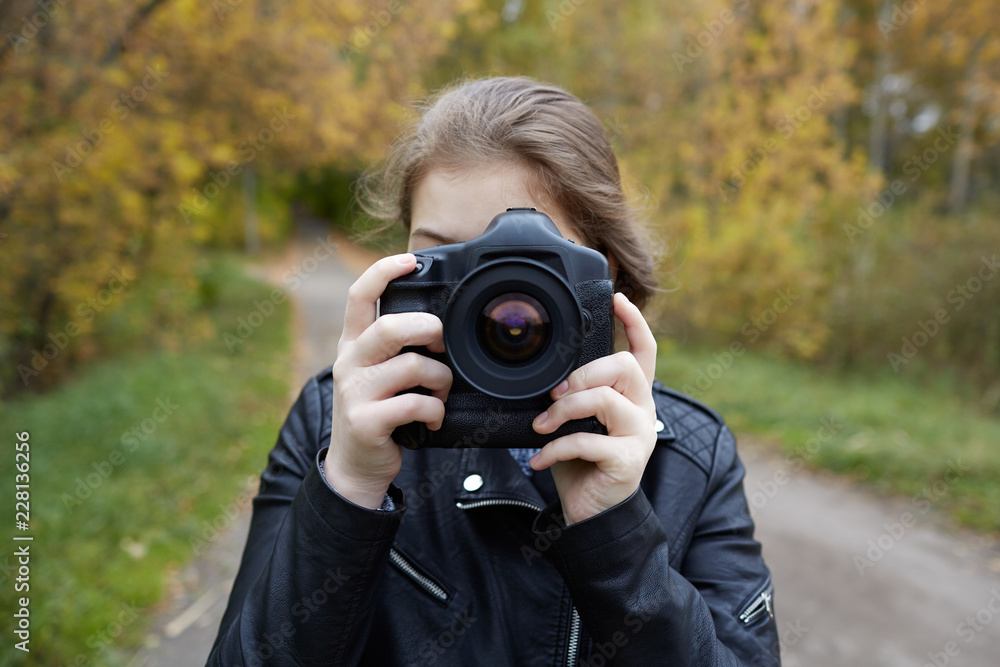 young girl in a leather jacket looks into SLR camera, ready to take a picture of a beautiful staged advertising, portrait, or reportage frame, against a blurred asphalt road, and autumn alley of trees young girl in a leather jacket looks into SLR camera, ready to take a picture of a beautiful staged advertising, portrait, or reportage frame, against a blurred asphalt road, and autumn alley of trees