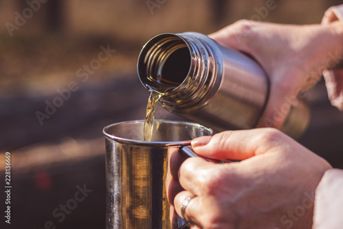 Woman pours hot tea out of thermos into metal cup in autumn forest photo