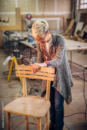 Female carpenter using drill to make a chair. Workshop interior.