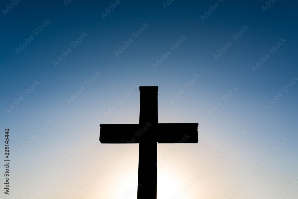 Simple oak wood catholic cross silhouette at sunset, dramatic dark blue sky with the glowing sun.