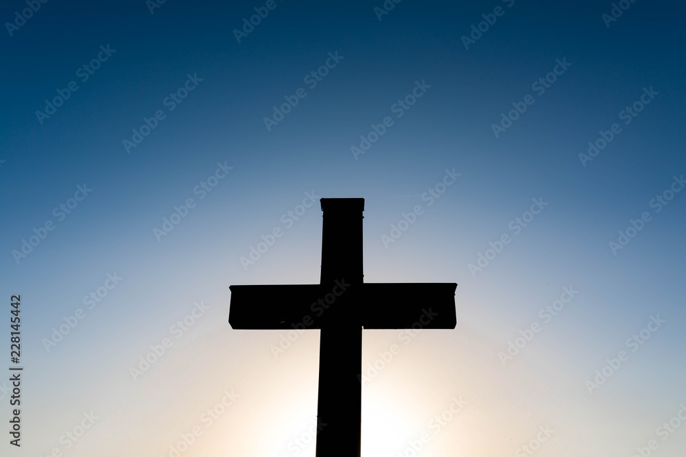 Simple oak wood catholic cross silhouette at sunset, dramatic dark blue sky with the glowing sun. Simple oak wood catholic cross silhouette at sunset, dramatic dark blue sky with the glowing sun.