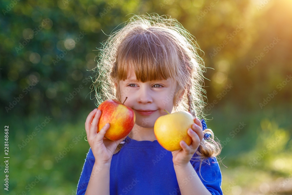 A cute smiling little girl holding ripe and juicy apples at the bright and sunny summer background. A cute smiling little girl holding ripe and juicy apples at the bright and sunny summer background.
