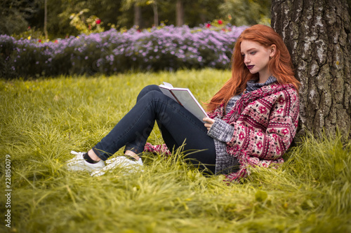 Redhead young concentrated woman with freckles and dark lipstick reading book outdoors in park sit under the big tree on grass with flowers on the background.