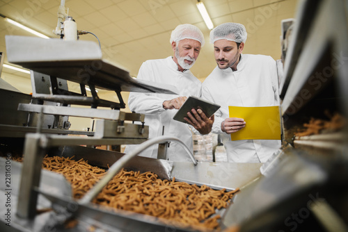 Two business man in sterile clothes standing in food factory in front of the production line and looking and tablet. Checking the quality of products and talking. photo