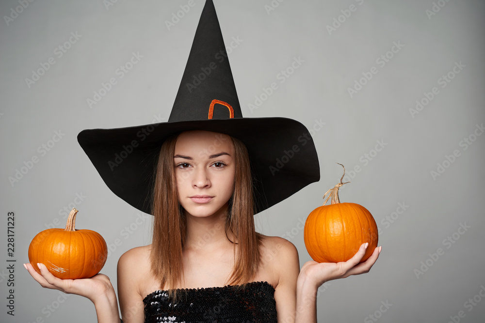 Closeup of serious Halloween witch holding orange pumpkins looking at camera with a look of suspect, isolated on grey background Closeup of serious Halloween witch holding orange pumpkins looking at camera with a look of suspect, isolated on grey background