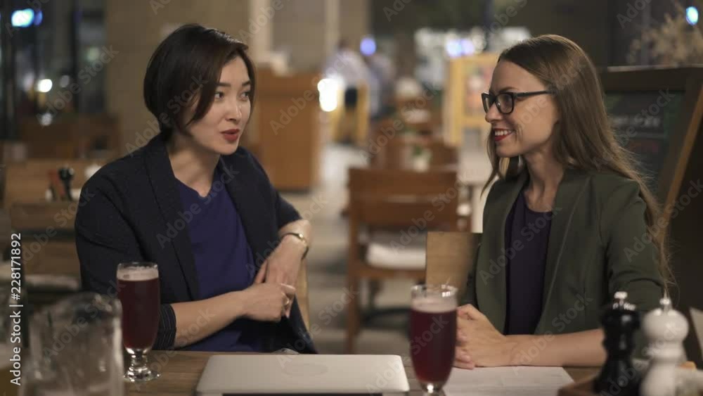 Attractive Asian woman with short hair talking to her serious caucasian friend wearing glasses in outdoors cafe at night. Concept of friendship. Handheld medium shot Attractive Asian woman with short hair talking to her serious caucasian friend wearing glasses in outdoors cafe at night. Concept of friendship. Handheld medium shot