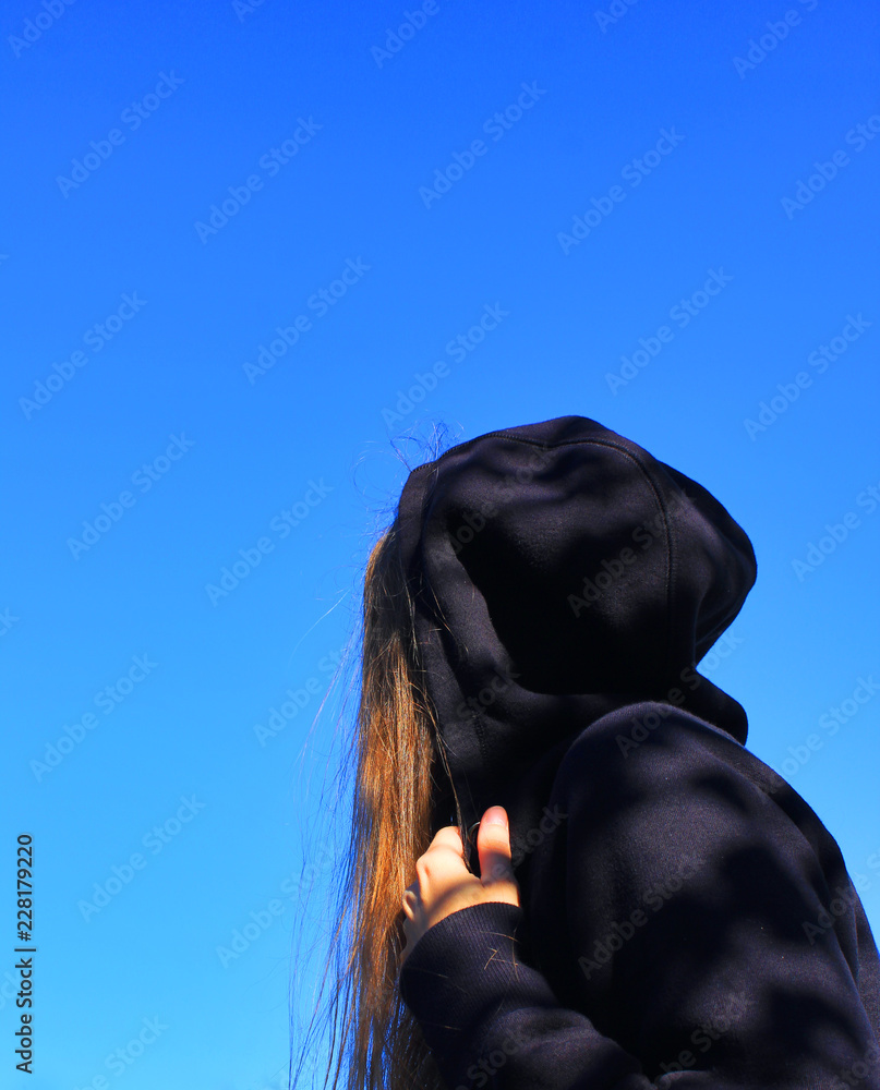 Young Woman Outdoors Wearing Hood on Black Casual Jacket. Young Stylish Girl Portrait on Empty Blue Sky Background. Day View of Lonely Female in All Black Clothes Standing Alone, Facing Sideways