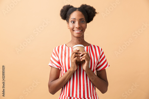 Happy african american woman in casual clothing holding paper cup with takeaway coffee, isolated over beige background