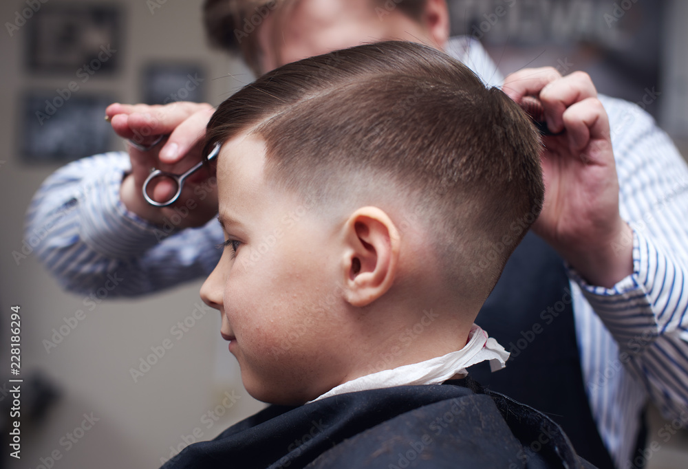 Fototapeta premium Barber making hairstyle to a Caucasian boy using scissors and hairbrush.