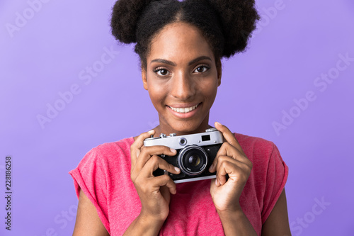 Photo of cheerful african american woman smiling and holding retro camera, isolated over violet background © Drobot Dean