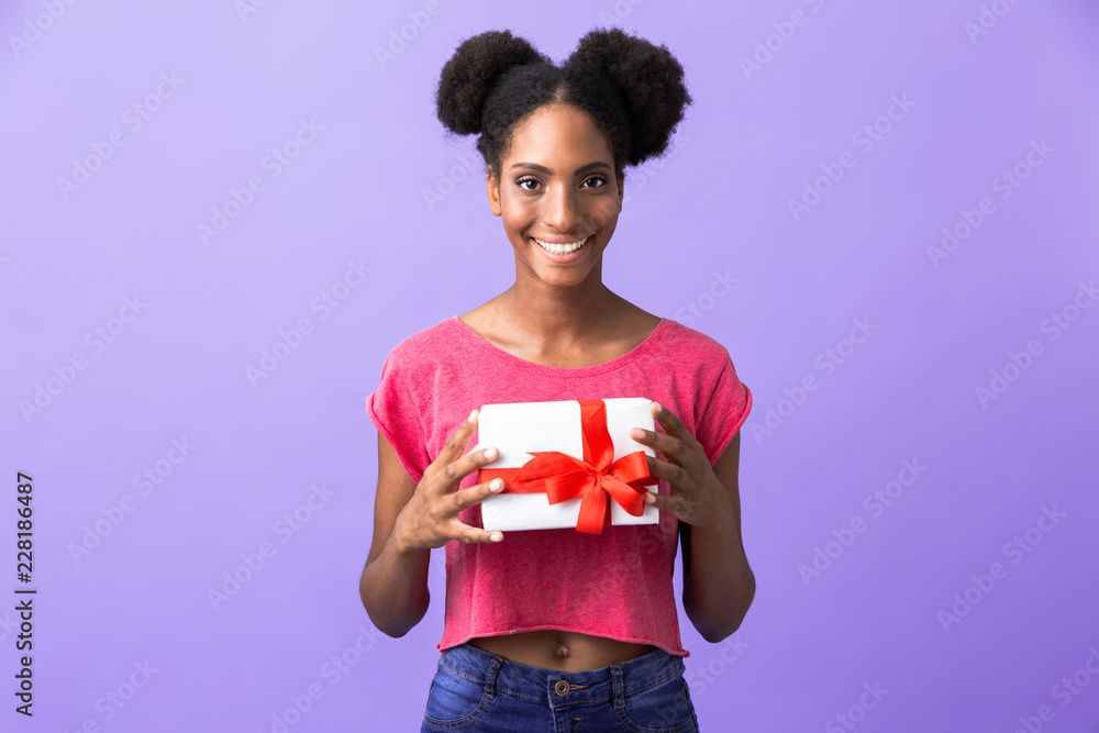 Photo of cute african american woman smiling and holding present box with red bow, isolated over violet background Photo of cute african american woman smiling and holding present box with red bow, isolated over violet background