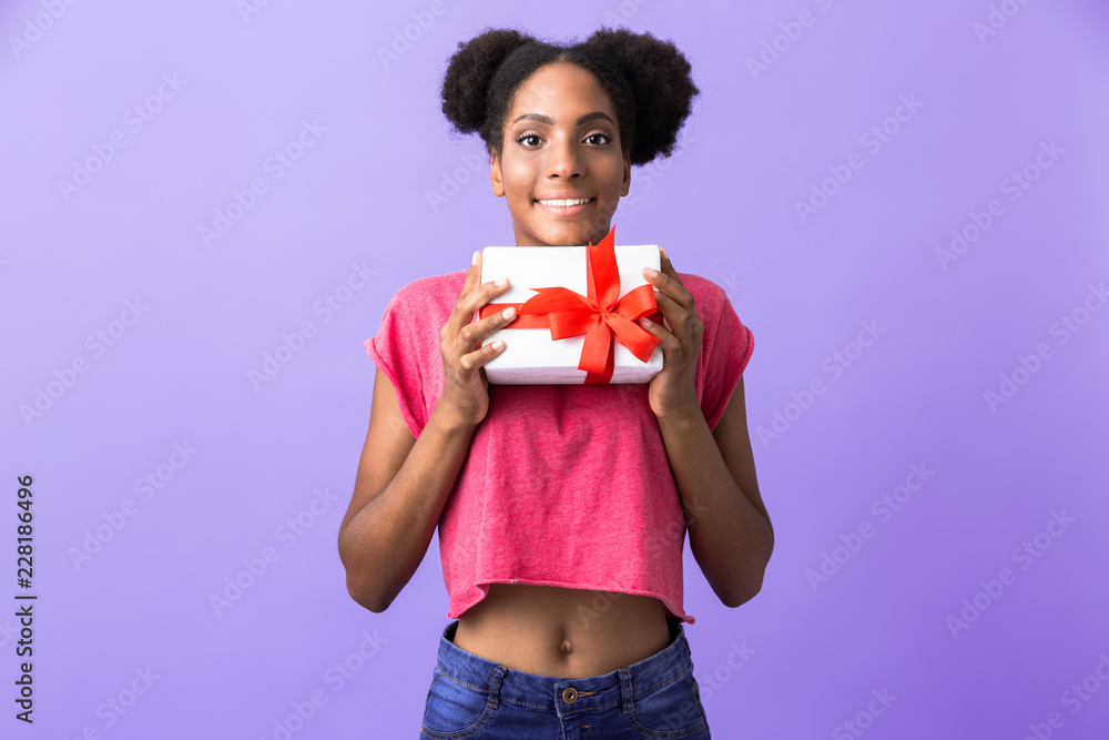 Photo of positive african american woman smiling and holding gift box with red bow, isolated over violet background Photo of positive african american woman smiling and holding gift box with red bow, isolated over violet background