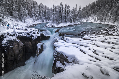 Cold scenery at Sunwapta Falls in the Canadian Rockies with snow and fir trees in wonderful scenery and ice and blue rivers running through the scenery with snowy hut