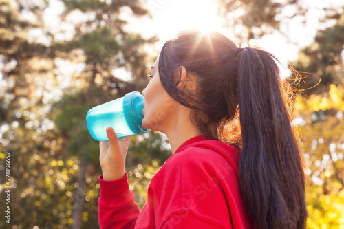 Athletic woman in sporty red clothes drinking water while jogging. Portrait, close-up in the sunset sunlight.